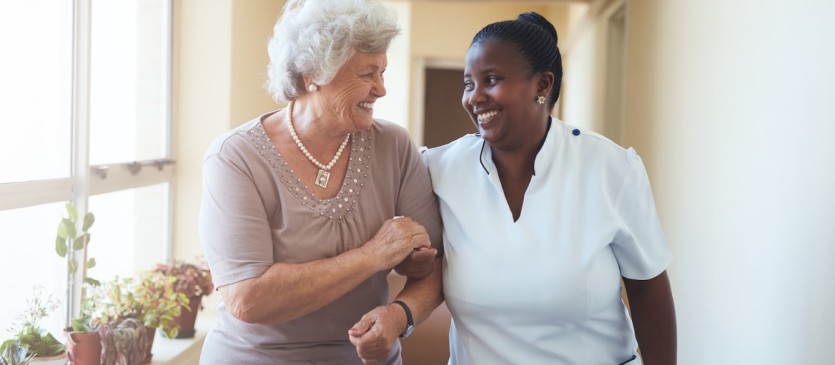 Portrait of smiling home caregiver and senior woman walking together through a corridor. Healthcare worker taking care of elderly woman.